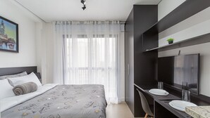 A modern hotel room with a bed, a desk with a chair, a TV, and a window with sheer curtains. - SB Setin Brooklyn Studios (São Paulo)