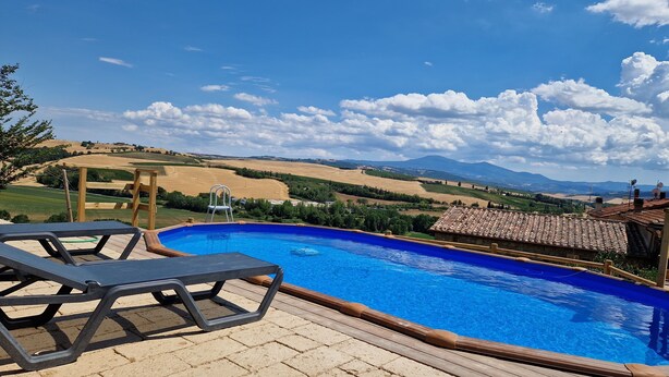 Una piscina con cubierta de madera y sillas de descanso, rodeada de un paisaje pintoresco con colinas y cielo despejado.