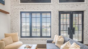 Una sala de estar con pared de ladrillo blanco, dos ventanas con marcos negros, un sofá color beige, un sofá gris y una mesa de centro de madera con un tablero de ajedrez.