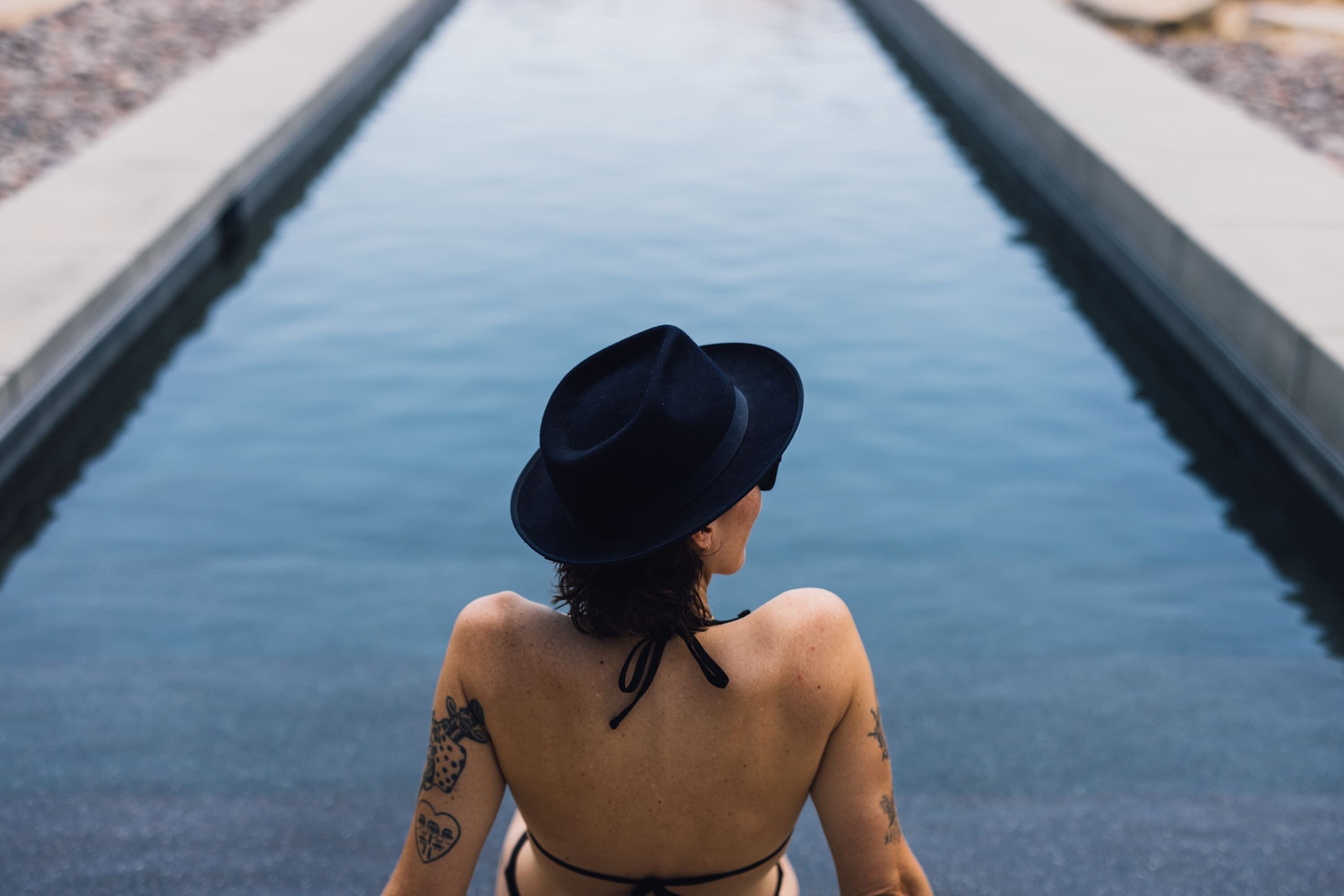 Une personne assise au bord d’une piscine, portant un chapeau, avec des tatouages dans le dos, et l’eau est calme.