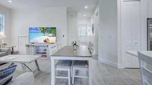 A modern kitchen with white cabinetry, a dining table with chairs, and a television displaying a beach scene. - Trendy Naples Retreat: Modern Vacation Home (Naples)