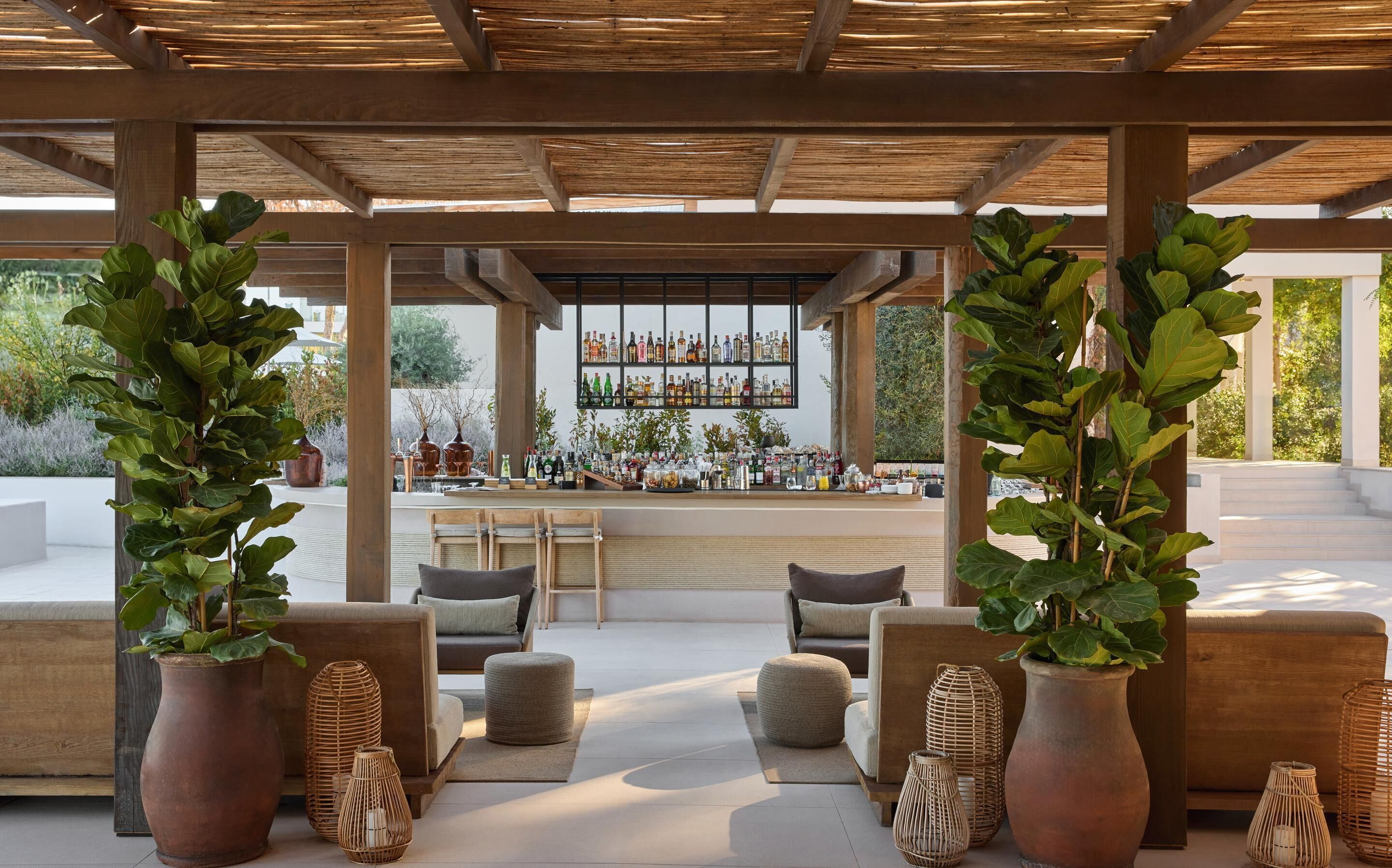 Un espacio al aire libre con techo y asientos, donde se destaca un bar al fondo, plantas en macetas y muebles de mimbre.