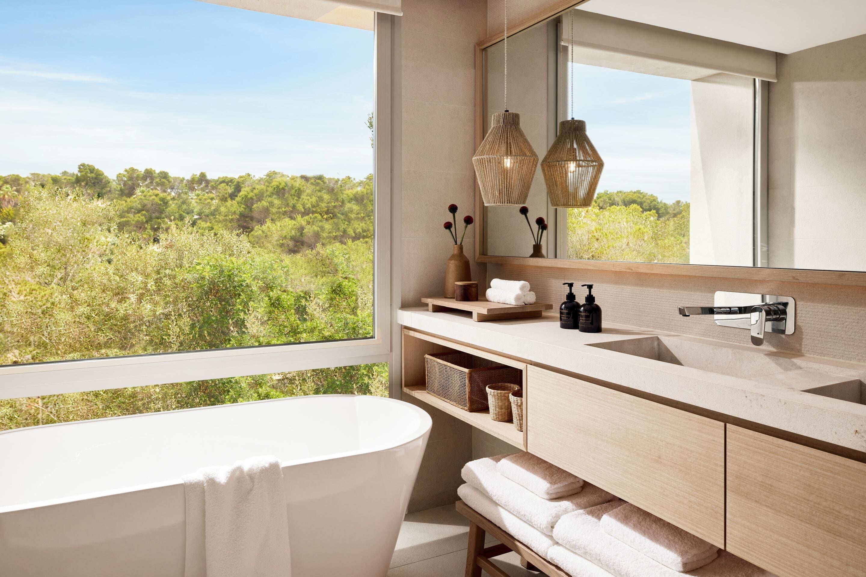 Ein modernes Badezimmer mit freistehender Badewanne, einem großen Spiegel und Blick auf eine üppige Landschaft.