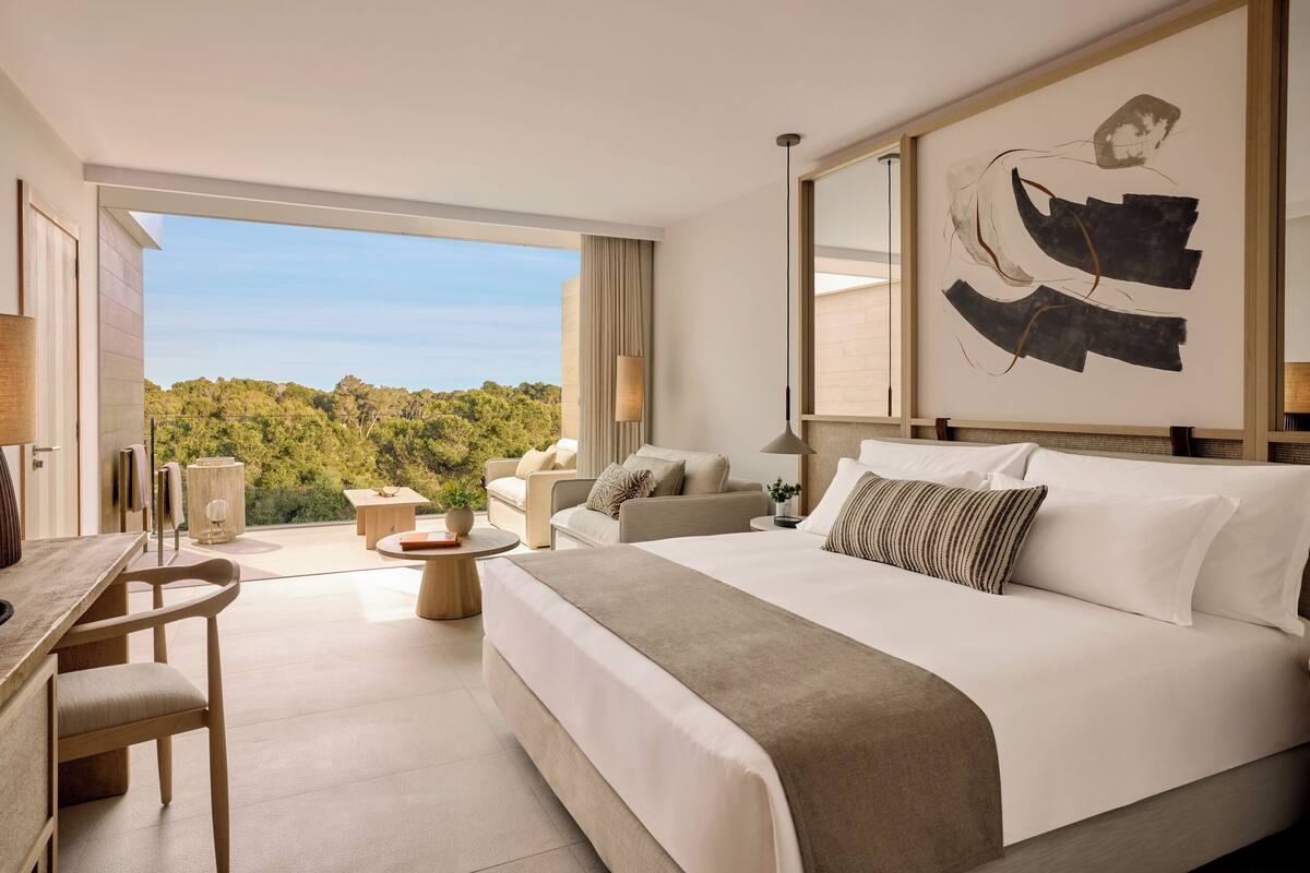 A modern hotel room with a large bed, a desk with a lamp, a chair, a small table, and a view of the outdoors.