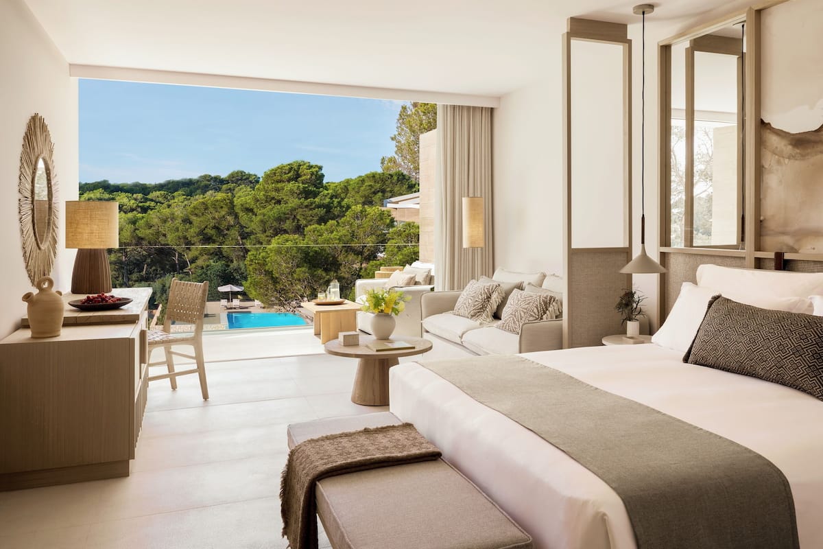 A modern hotel room with a large bed, a sofa, a desk, and a view of a pool and greenery.