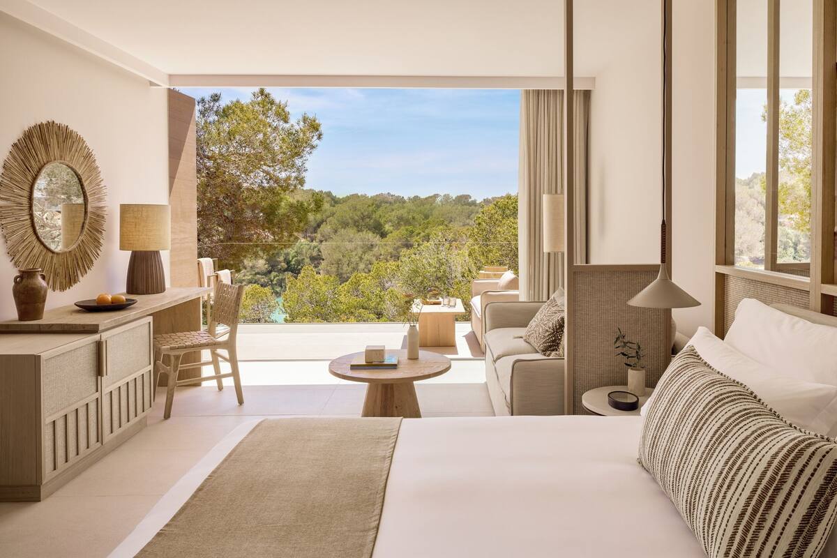 A modern hotel room with a large bed, a sitting area with a sofa and chairs, a small round table, and a balcony with a view of the forest.