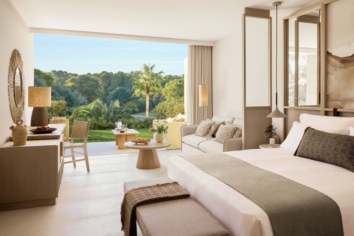 A hotel room with a large bed, a sofa, a dining table, and a view of a lush garden.