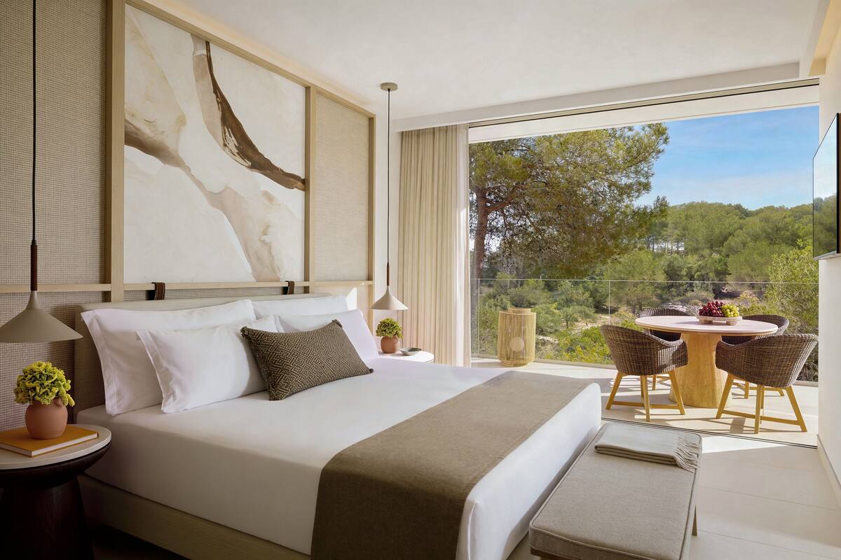 A modern hotel room with a large bed, a dining area with wicker chairs, and a balcony with a view of trees.