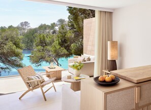 A modern hotel room with a balcony overlooking a turquoise sea, a wooden desk, a sofa, and a lamp. - Ikos Porto Petro – All inclusive (Santanyi)
