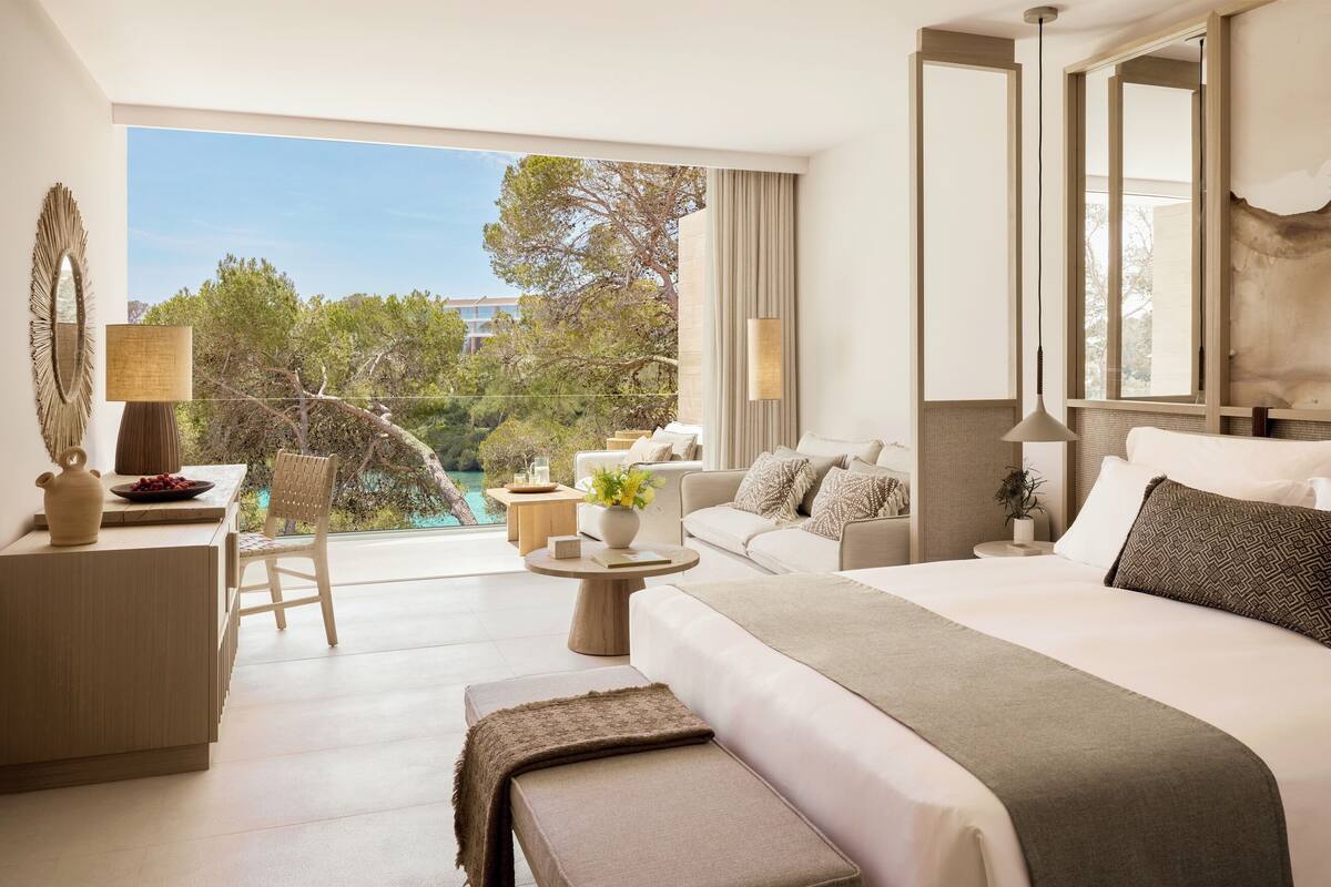A modern hotel room with a large bed, a sofa, a dining table, and a balcony with a view of trees and a body of water.