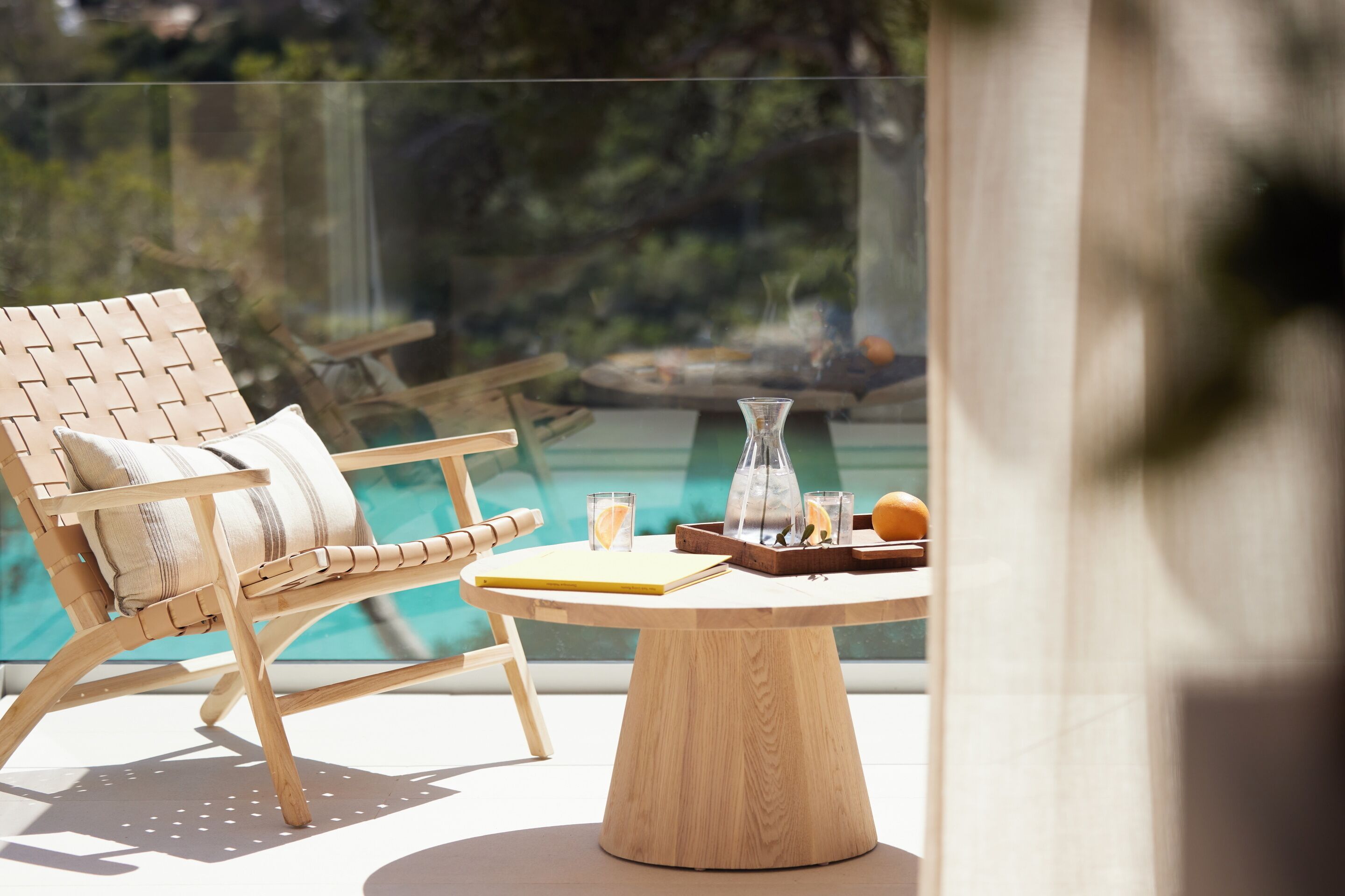 A wooden lounge chair with a cushion, a round wooden table with a tray, and a glass vase.