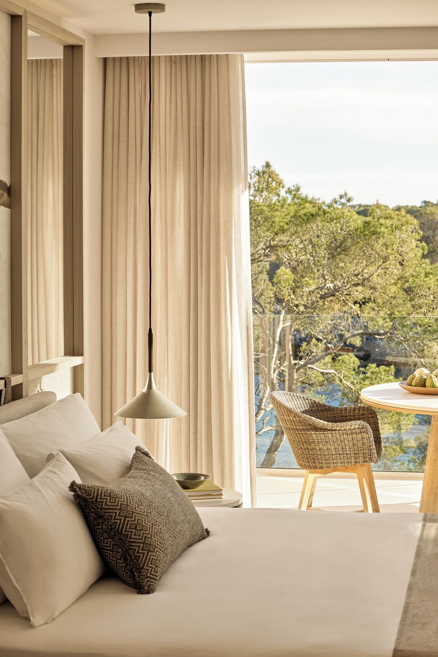 Un dormitorio con cama, almohadas, lámpara colgante, una silla y vistas a árboles y agua a través de una ventana.