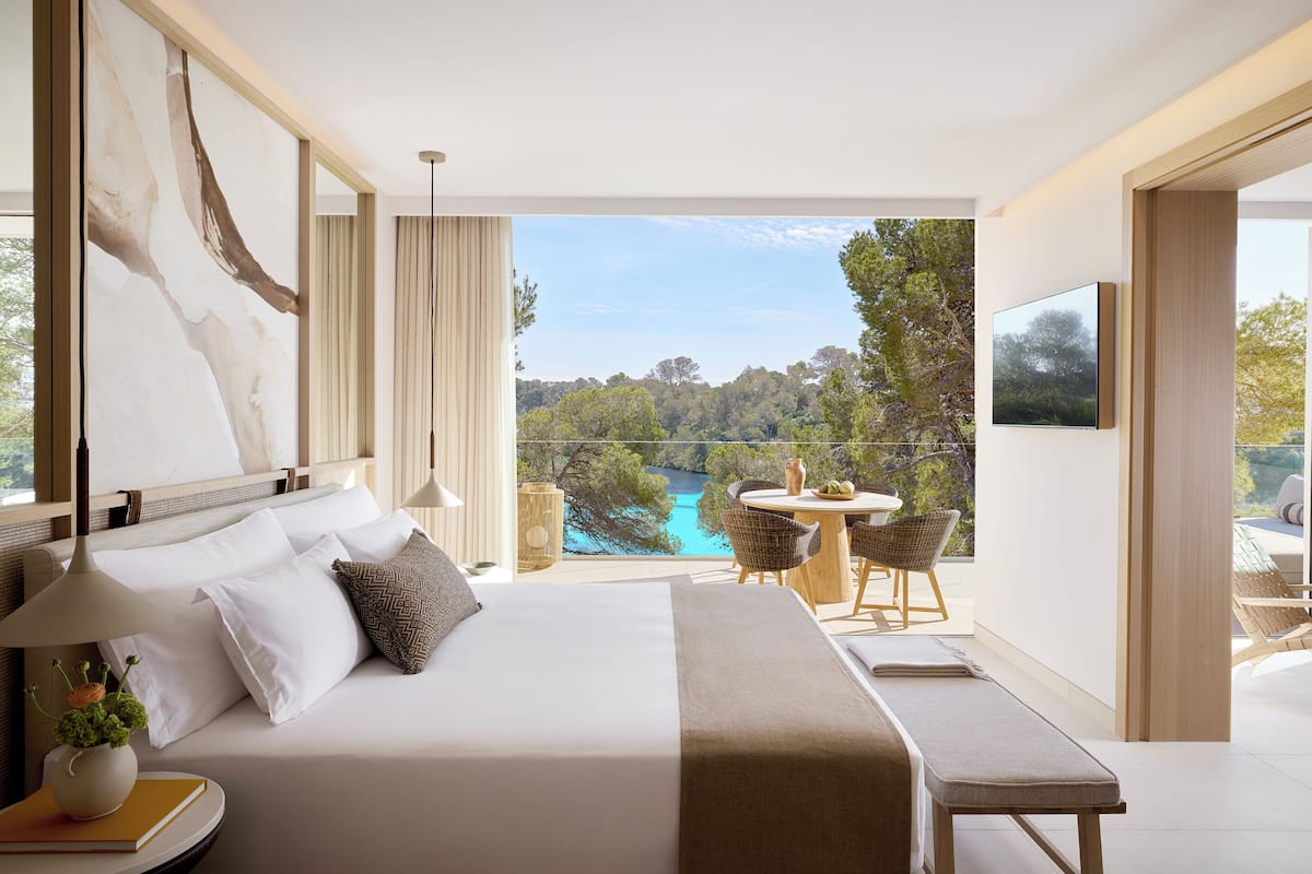 A modern hotel room with a large bed, a bedside table with a lamp, a view of a lake, and a balcony with outdoor seating.