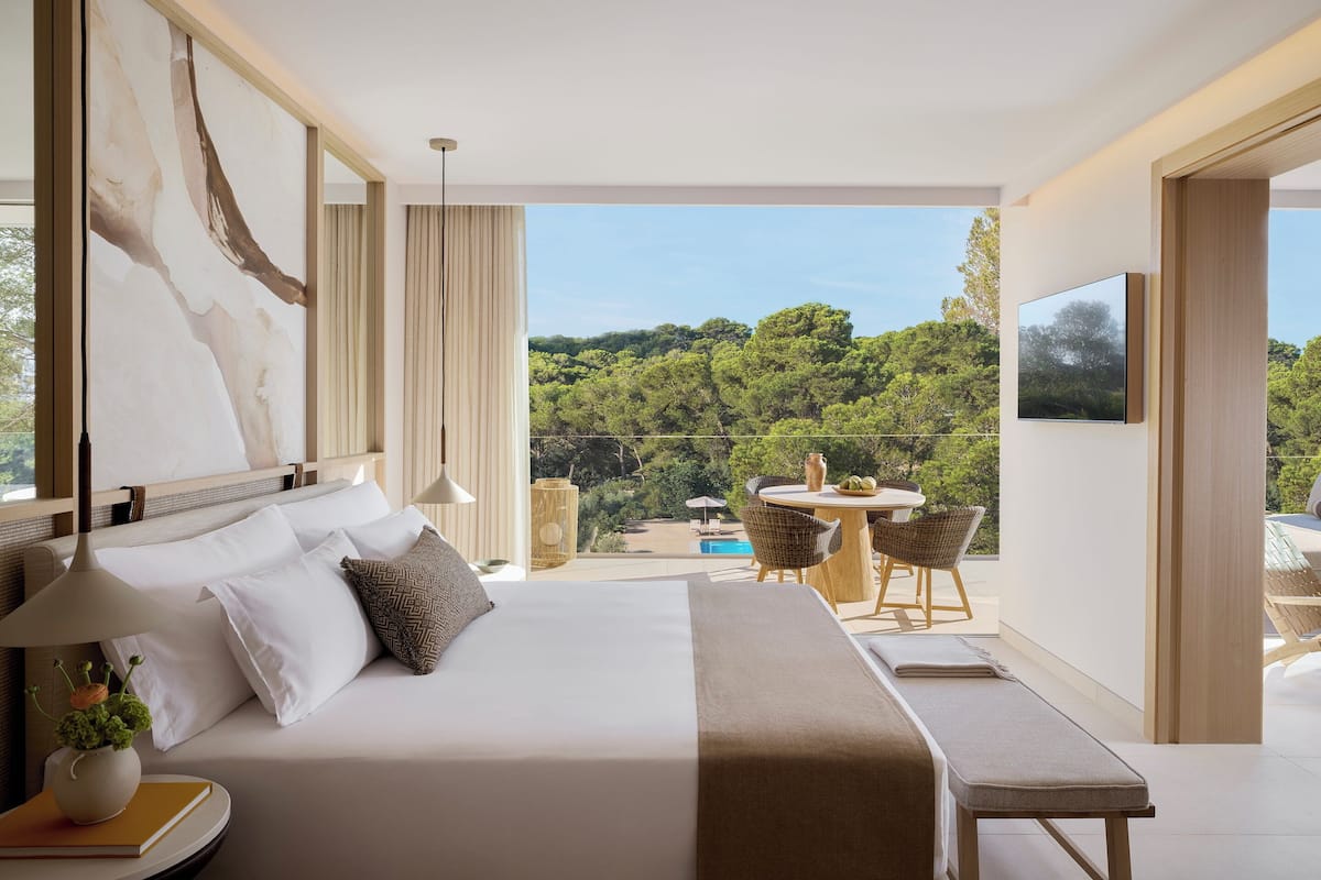 A modern hotel room with a large bed, a balcony with a table and chairs, and a view of a lush green landscape.