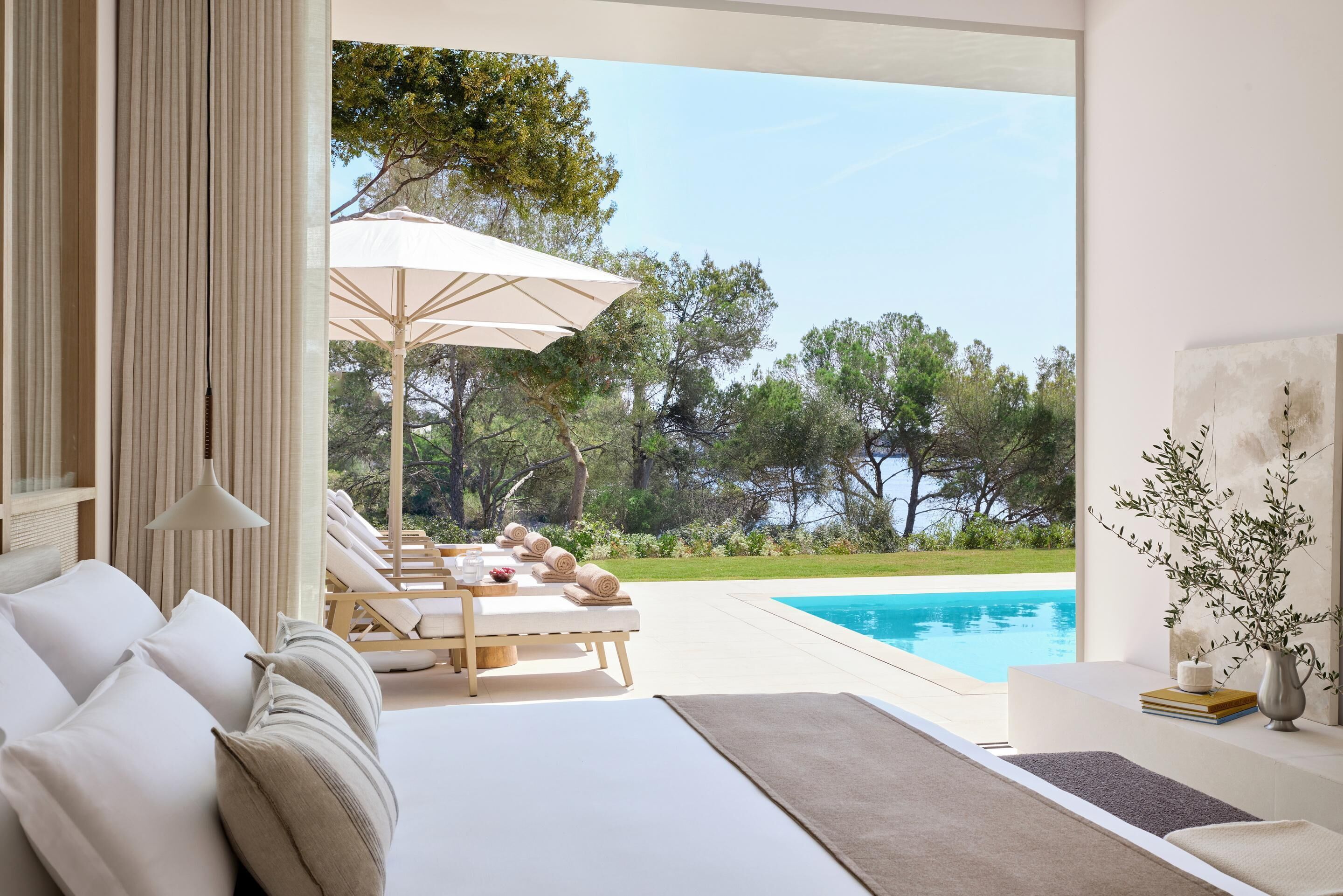 A bedroom with a large bed, a view of a swimming pool, and a sun lounger outside.