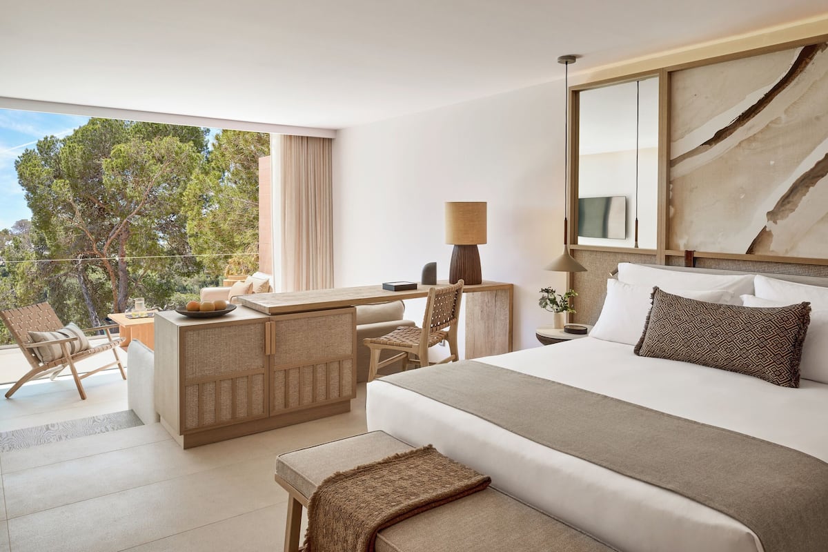 A hotel room with a large bed, a desk, a chair, a lamp, and a view of trees.