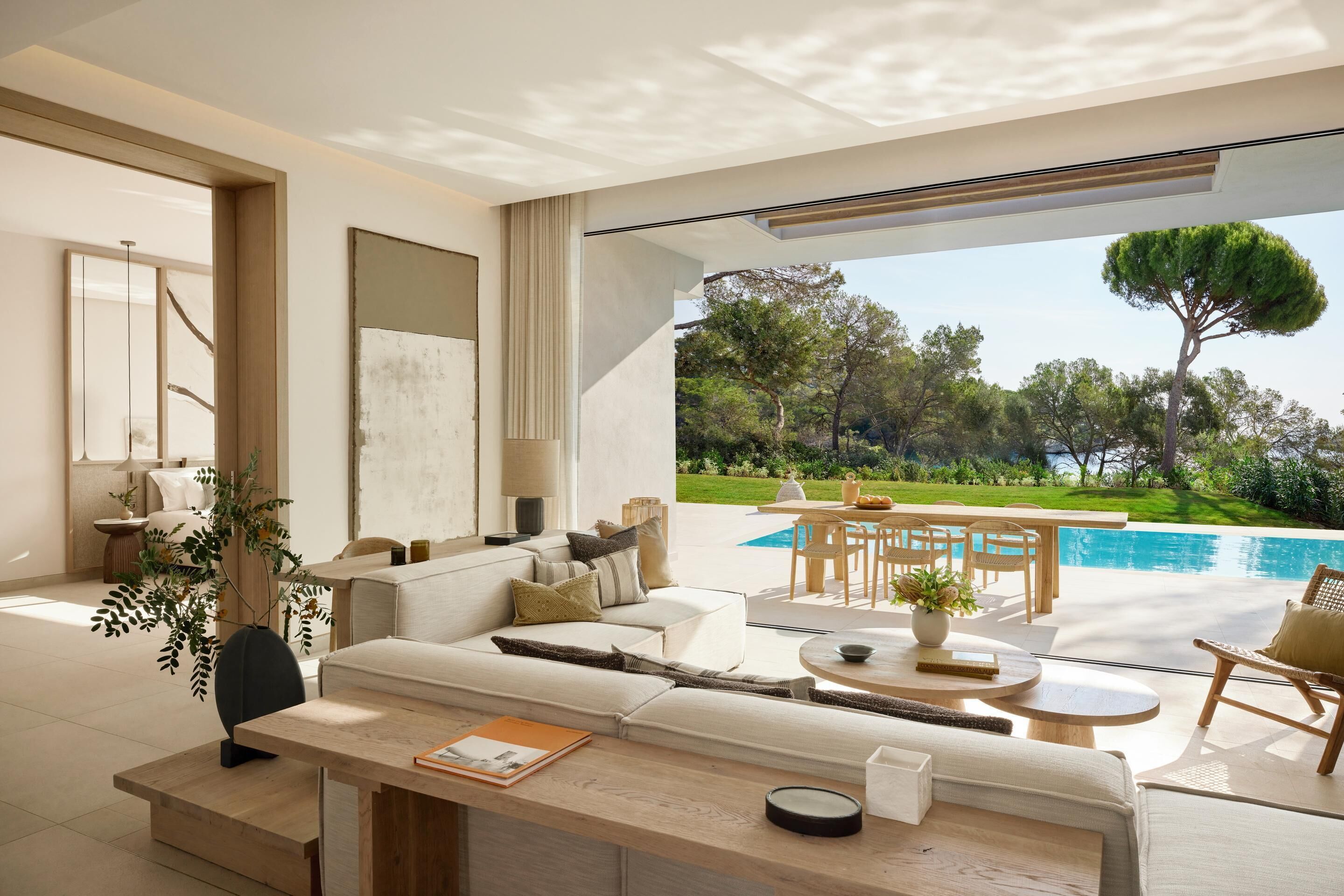 A modern living room with a sofa, coffee table, and dining area, overlooking a swimming pool and lush greenery.