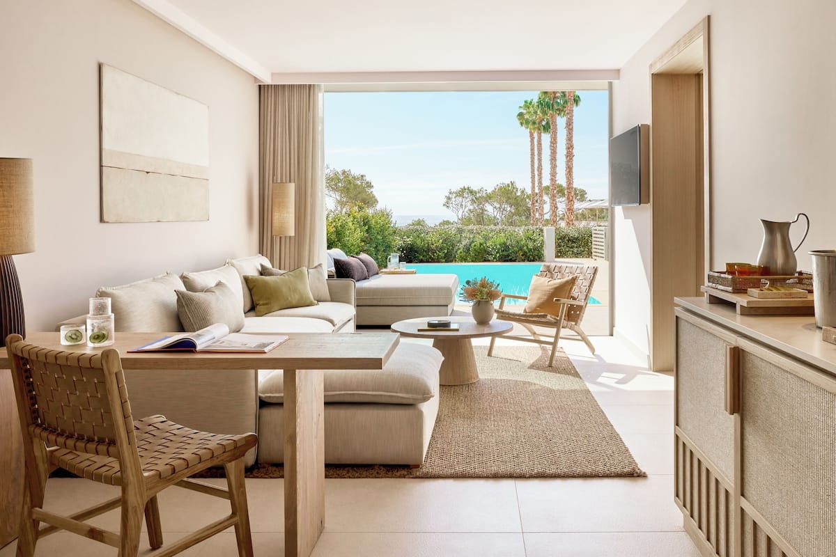 A modern living room with a sofa, coffee table, and chairs, a dining table, a TV, and a view of the ocean and palm trees.