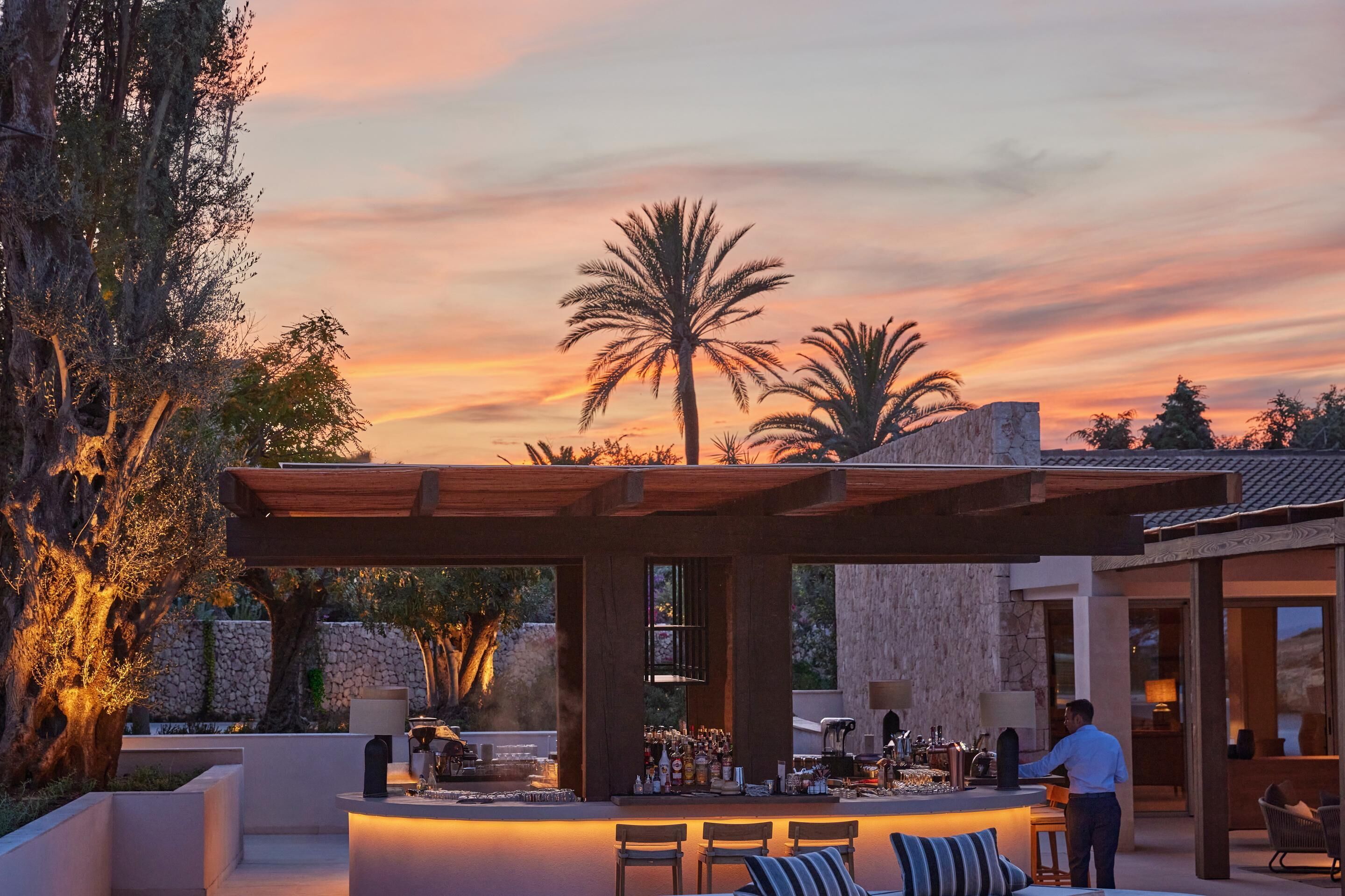 An outdoor bar area with seating, a stone wall, and palm trees.