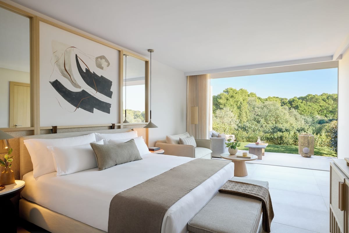 A modern hotel room with a large bed, a sofa, a small table, and a view of a forest.