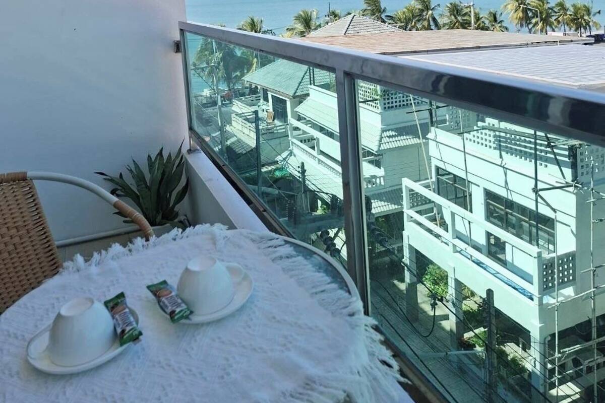 En balkon med udsigt over havet, et bord med kopper og en plante, samt en hvid væg med et klimaanlæg.