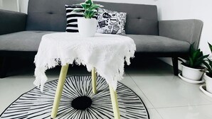 Ein modernes Wohnzimmer mit einem grauen Sofa, einem runden Teppich in Schwarz-Weiß-Muster, einem kleinen weißen Tisch mit einer verzierten Tischdecke und zwei gerahmten Wandbildern.