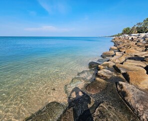 A rocky shoreline with clear water, a distant view of a building, and a clear sky. - The Sunset Bangsaen Seaview (Chonburi)