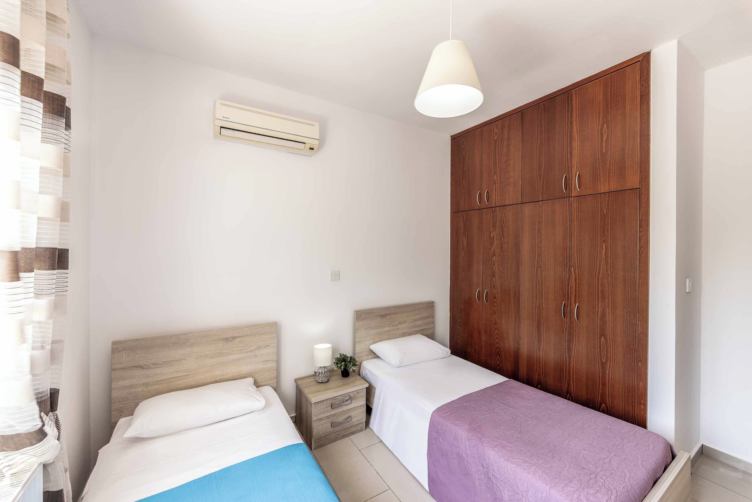 Une chambre avec deux lits, une armoire en bois, une table de chevet avec une lampe et un appareil de climatisation.