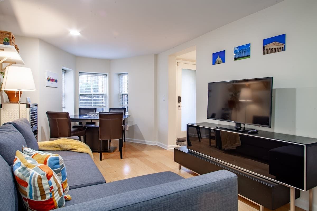 A modern living room with a grey sofa, a flat-screen TV, and a dining area with a table and chairs.