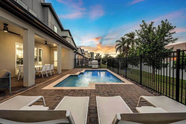 A modern outdoor pool area with lounge chairs, a barbecue grill, and a view of the sunset.