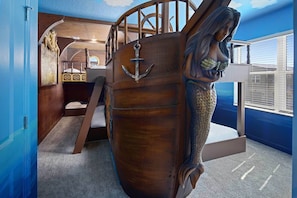A room with a large wooden ship's wheel, a bed with a white headboard, and a window with blinds.