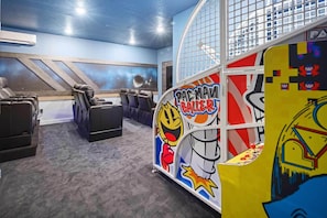 A room with a blue ceiling, a large window, and a wall mural of space. There are two black leather chairs and a Pac-Man arcade machine.