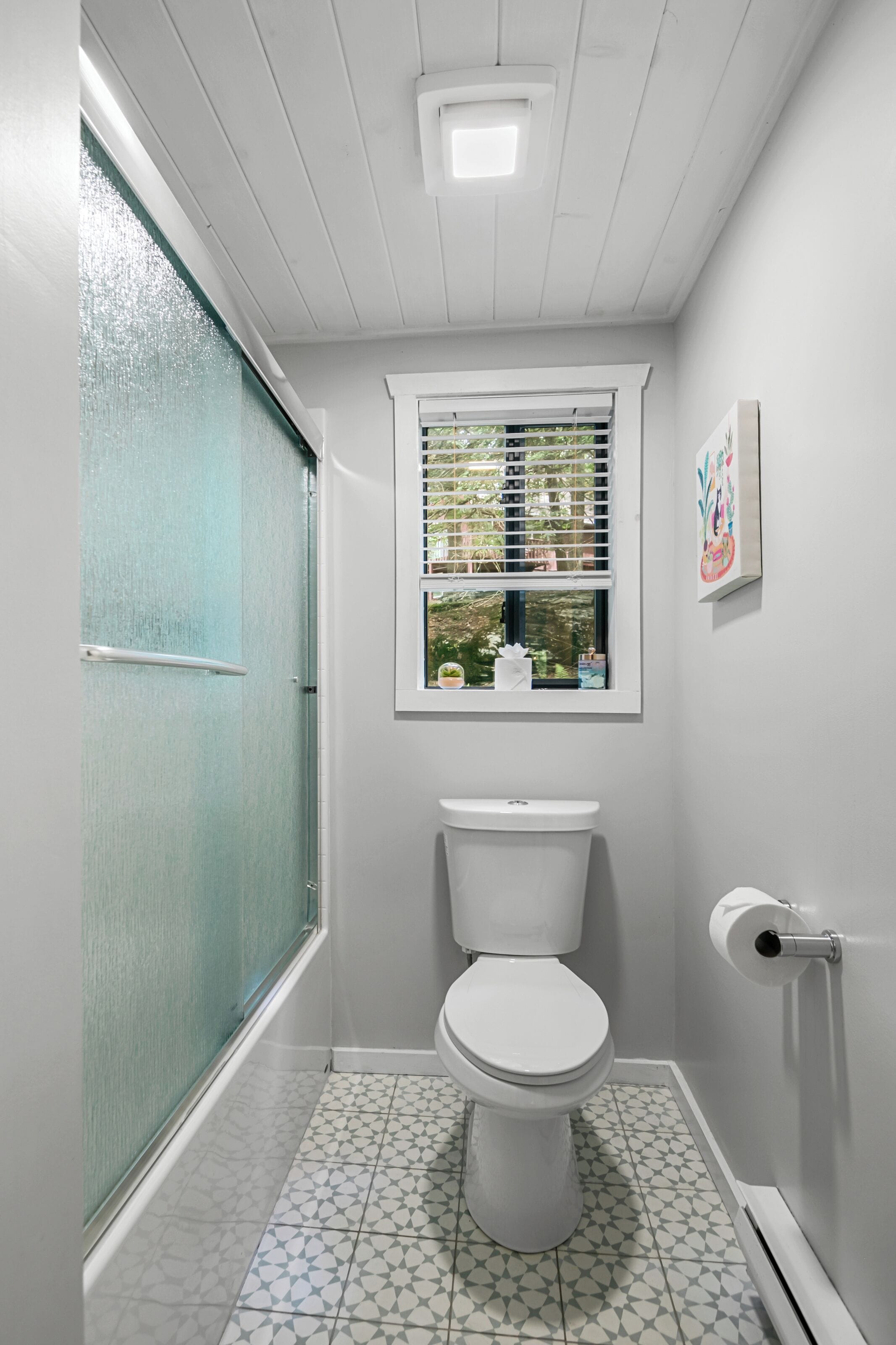 Een kleine badkamer met toilet, een raam met zonwering en een douchedeur.