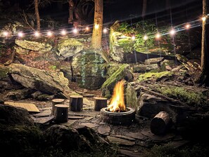 A stone fire pit surrounded by logs and rocks, with string lights above.
