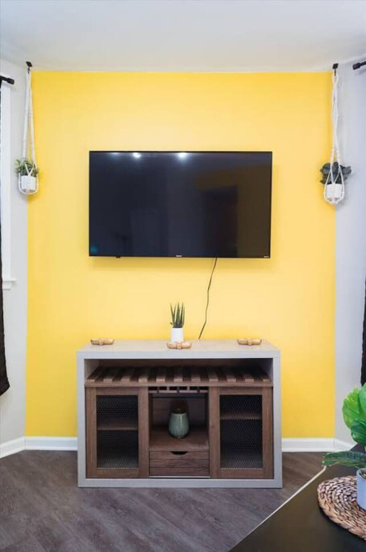 A room with a yellow wall, a mounted TV, a wooden console table with a plant, and a small cabinet.