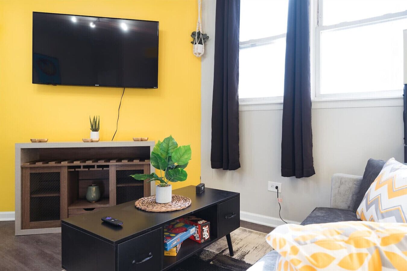 A room with a flat-screen TV mounted on a yellow wall, a black entertainment unit with a plant, a bed with a patterned blanket, and a window with dark curtains.