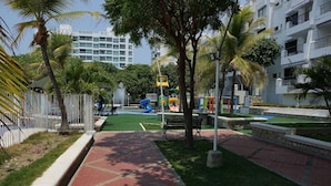 Un parque infantil con toboganes y un banco, rodeado de palmeras y una valla blanca.