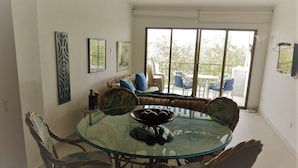 Un comedor con una mesa redonda de vidrio rodeada de sillas de mimbre. Hay cuadros en la pared y una ventana con vista a muebles de exterior.