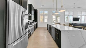 A modern kitchen with a stainless steel refrigerator, black cabinets, and a marble countertop. There are pendant lights, a built-in oven, and a large window providing natural light. - Sunrise A🌞Waterfront👙Pool & Hot Tub🐶Dogs OK (Okaloosa Island)