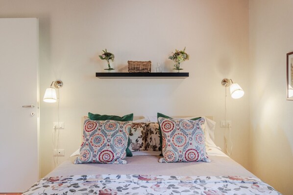 Un dormitorio con una cama, dos lámparas fijadas a la pared, un estante con adornos y una puerta.