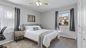 Un dormitorio con cama, mesitas de noche, escritorio, silla, ventilador de techo y ventana con cortinas.