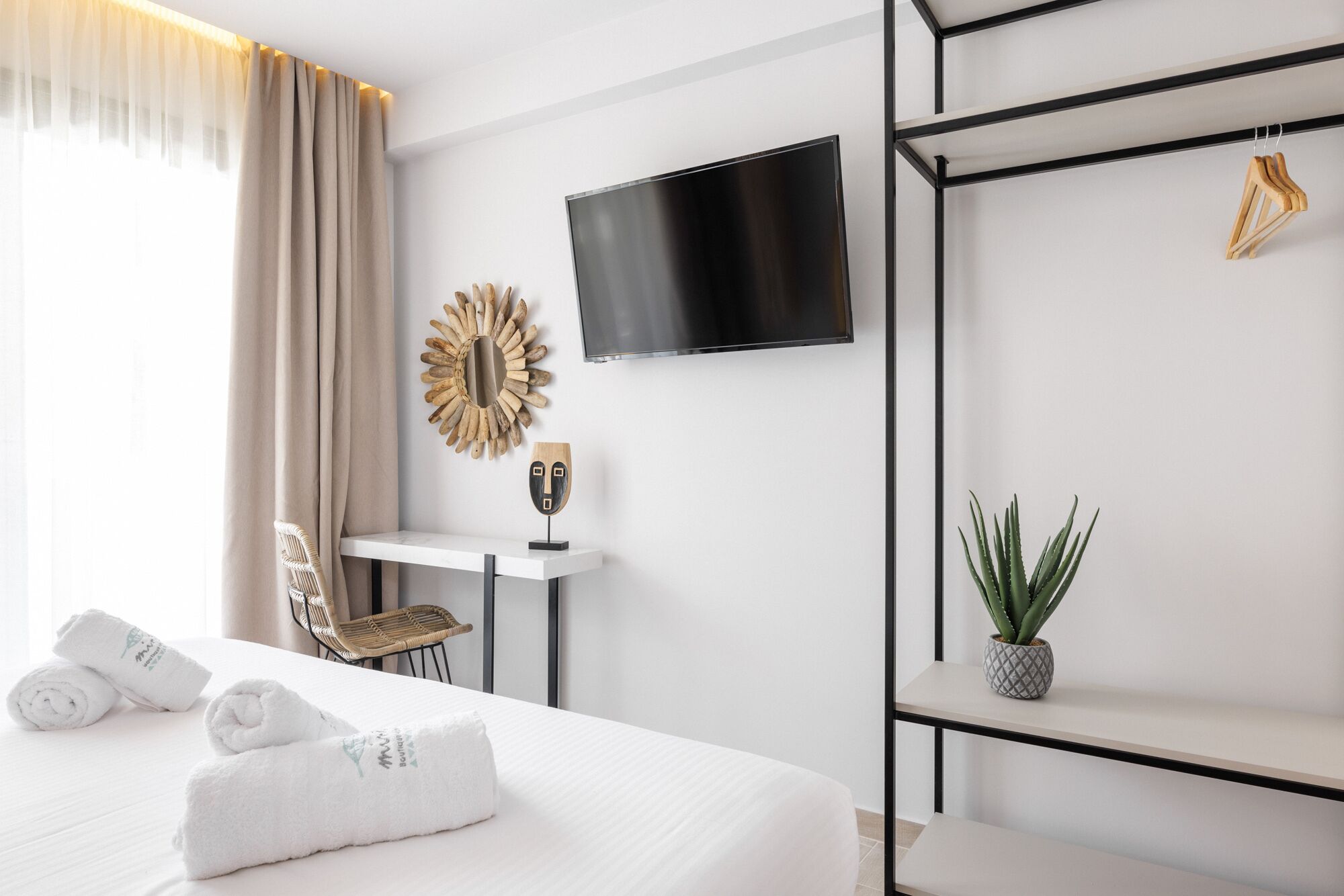 A hotel room with a bed, a TV mounted on the wall, a small table with a mirror, a chair, and a potted plant.
