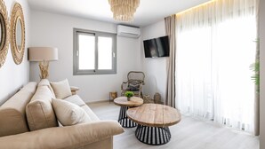 A modern living room with a sofa, a round wooden coffee table, a wicker chair, a TV mounted on the wall, and a large window with sheer curtains. - Amalthia Luxury Suites (Kassandra)