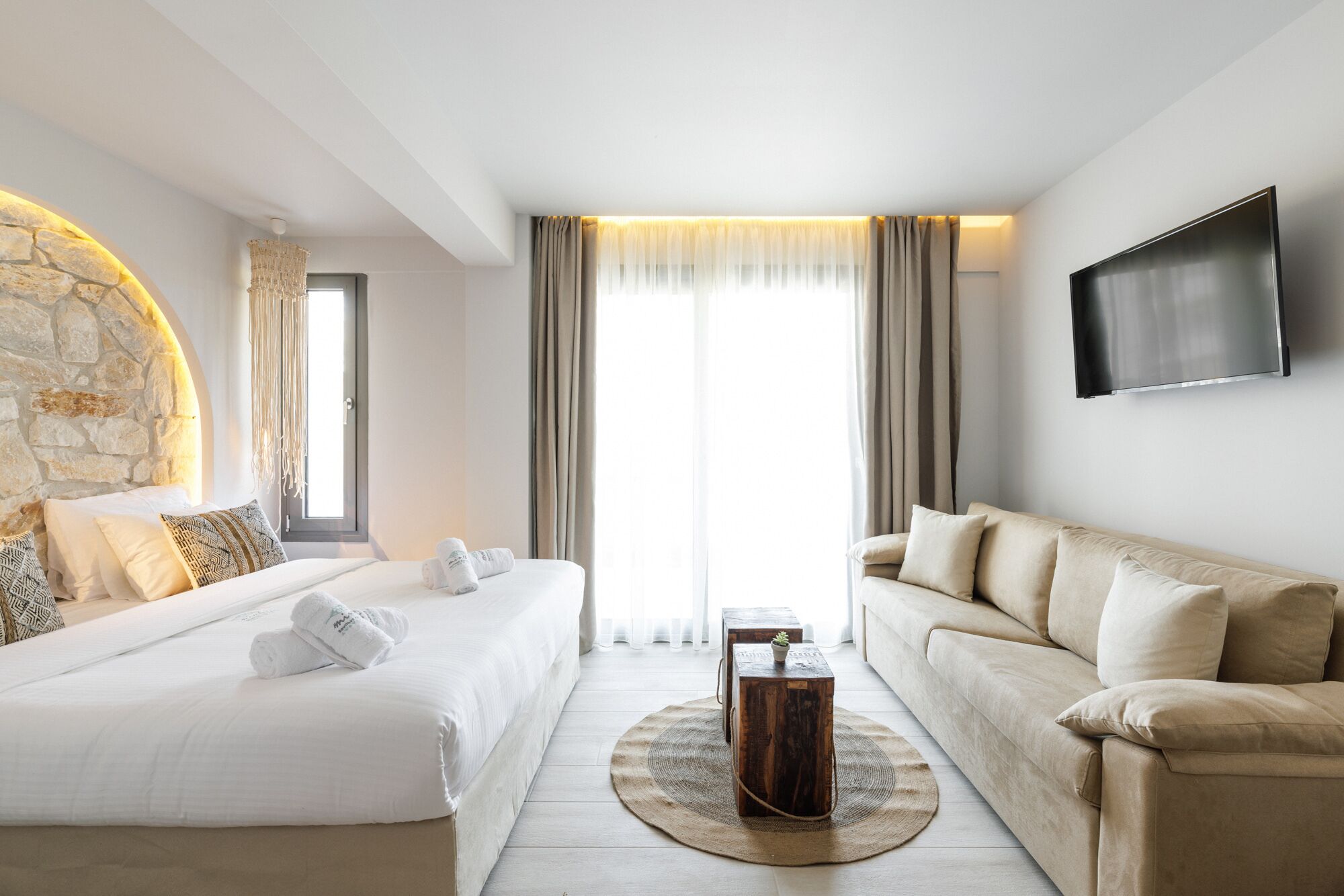 Ein modernes Hotelzimmer mit einer Wand aus Stein, einem Bett mit weißen Leinentextilien, einem beigen Sofa, einem runden Teppich und einem an der Wand montierten Flachbildfernseher.