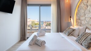 A hotel room with a bed, a TV, a stone accent wall, a view of the sea, and a balcony with chairs. - Amalthia Luxury Suites (Kassandra)
