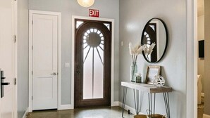 Un couloir avec une porte blanche, un panneau de signalisation indiquant la sortie, une porte en bois dotée d’une fenêtre ronde, un miroir et une console ornée d’objets décoratifs.