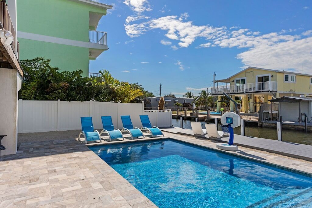 A swimming pool with blue chairs, a basketball hoop, and a view of a water body and buildings.