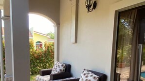 A covered outdoor seating area with a sofa and table, a wall-mounted light, and a view of a yellow building and greenery. - Casa Dulce Vida Gran Pacifica Resort (Villa El Carmen)