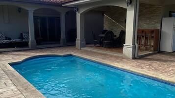 Piscina com deck azulejado, área de varanda coberta com cadeiras e um edifício com telhado de telhas vermelhas.
