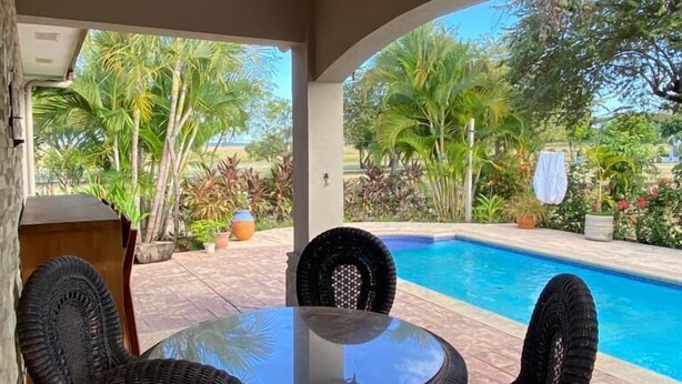 A covered patio with a glass table and wicker chairs, overlooking a swimming pool and lush greenery. - Casa Dulce Vida Gran Pacifica Resort (Villa El Carmen)